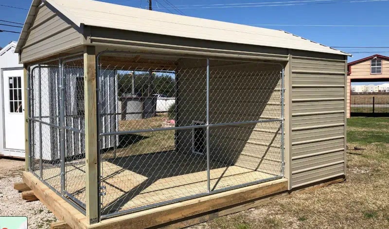 Tan metal portable building with a chain-link kennel, on a gravel lot with other sheds behind it.